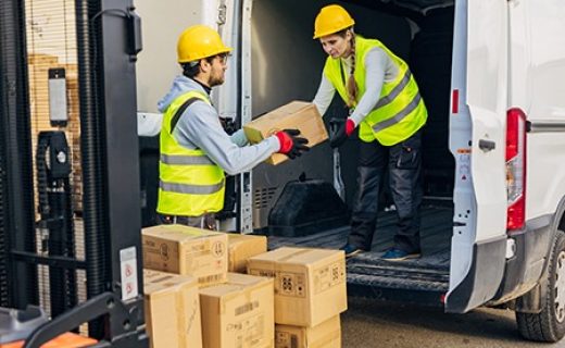 A male worker in a reflective uniform passes cardboard boxes to a female colleague standing in a van and arranges them and prepares them for delivery