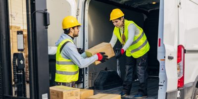 Two warehouse workers transfer cardboard boxes from a forklift to a delivery van A male worker in a reflective uniform passes cardboard boxes to a female colleague standing in a van and arranges them and prepares them for delivery