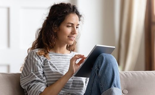 Smiling young woman using computer tablet, sitting on couch, typing on touchscreen, chatting in social network with friends, playing mobile device game or surfing internet, shopping online
